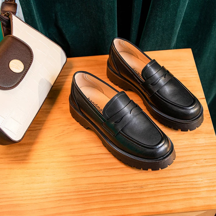 Black loafers on a wooden surface with a handbag in the background
