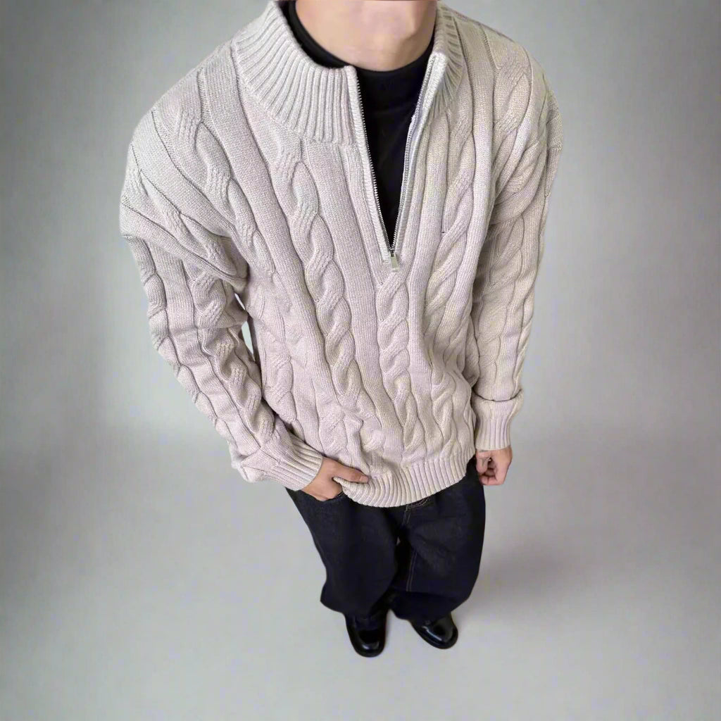 Person wearing a beige cable knit sweater over a black shirt on a plain background