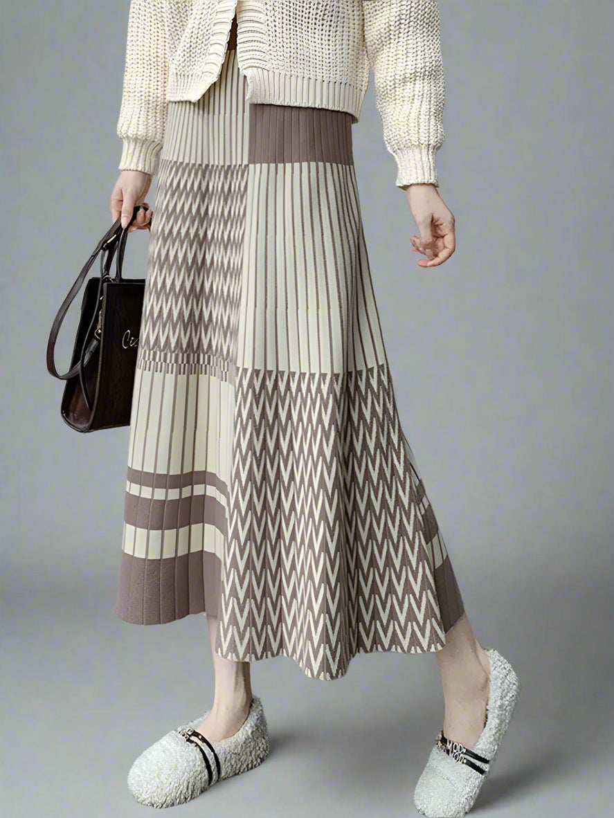 Person wearing a patterned skirt with a neutral sweater and shoes, holding a black handbag.