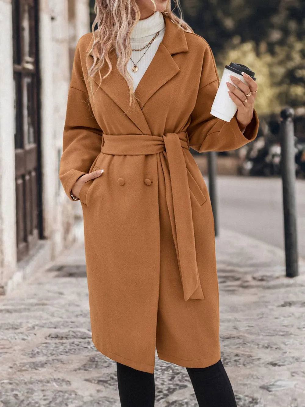 A woman wearing a caramel coat holding a white cup 