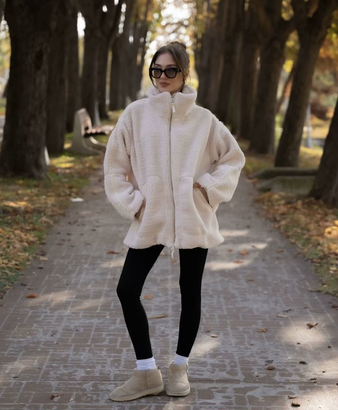 Person wearing a white fleece jacket and black leggings standing on a path with trees in the background.
