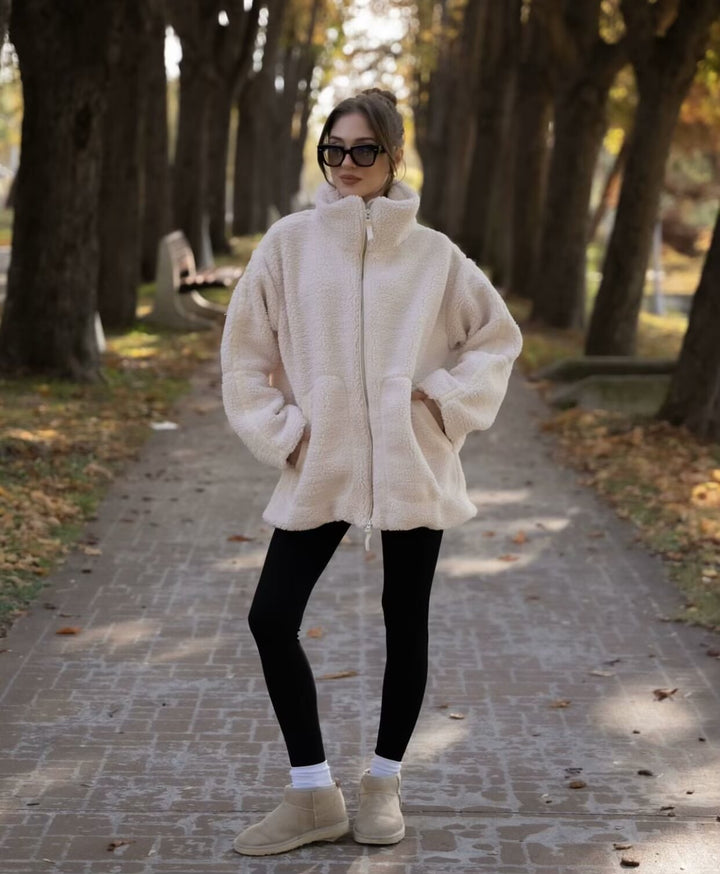 Person wearing a white fleece jacket and black leggings standing on a path with trees in the background.