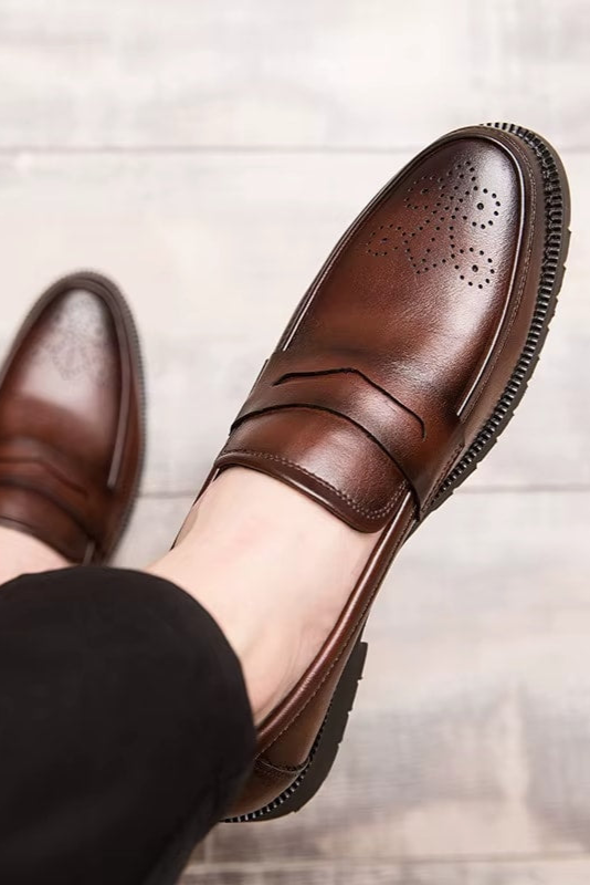 Brown leather shoes worn with black pants on a light wooden floor.