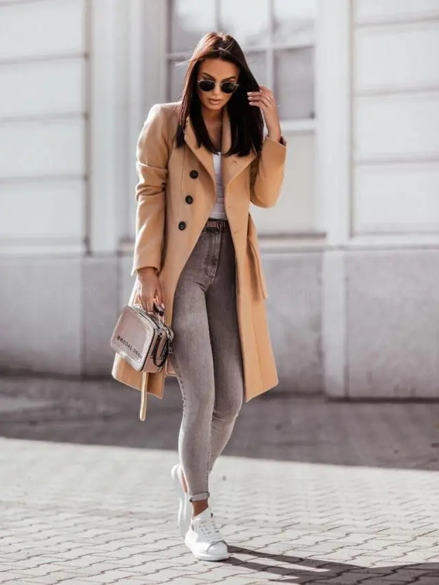 Woman wearing a beige coat, gray jeans, and white sneakers on a city street.