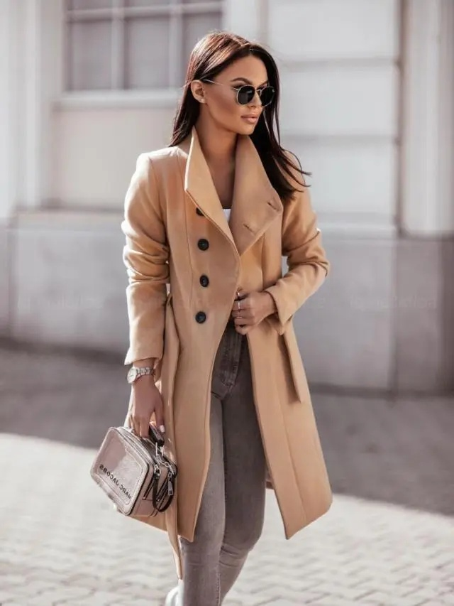Woman wearing a beige coat and sunglasses, holding a handbag on a street.